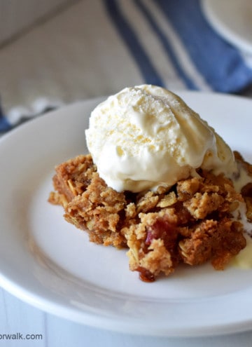 Close up view of a serving of gluten free rhubarb crumble, topped with vanilla ice cream, on a small white dessert plate with a blue and white striped towel and more crumble in the background.