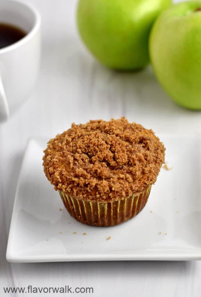 One gluten free apple muffin on a small white plate with a cup of coffee and two granny smith apples in the background.