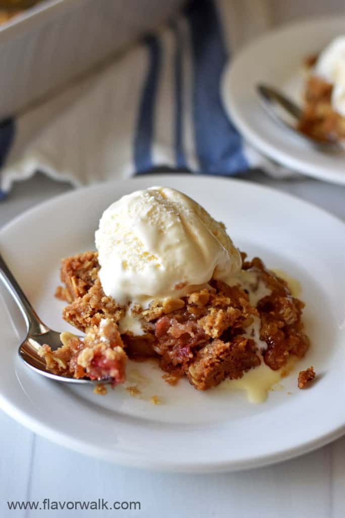 Close up view of a serving of gluten free rhubarb crumble topped with vanilla ice cream on a small white plate with a spoon and more rhubarb crumble in the background.