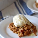 Close up view of a serving of gluten free rhubarb crumble topped with a scoop of vanilla ice cream on a white dessert plate with a blue and white striped towel and more rhubarb crumble in the background.