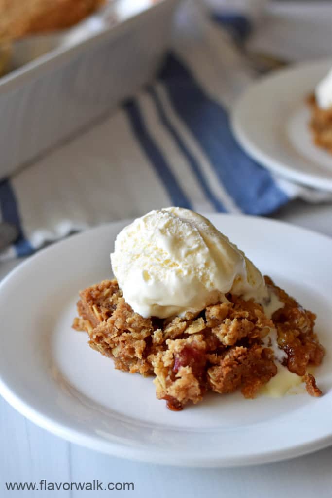 Close up view of a serving of gluten free rhubarb crumble topped with a scoop of vanilla ice cream on a white dessert plate with a blue and white striped towel and more rhubarb crumble in the background.