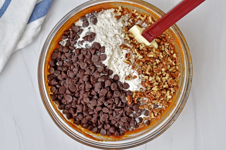 Overhead view of glass mixing bowl with chocolate chips, gluten free flour blend, and chopped pecans being added to batter for gluten free chocolate chip pumpkin bread with a rubber spatula. A blue and white striped kitchen towel is in the upper left corner.