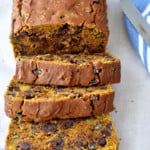 Close up view of 3 slices of gf chocolate chip pumpkin bread and the remaining loaf on parchment paper with a bread knife and blue and white striped kitchen towel on the right.