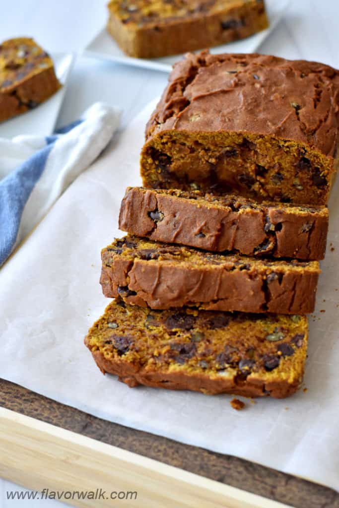 A sliced loaf of gluten free chocolate chip pumpkin bread on parchment paper with a blue and white striped kitchen towel and more slices on white plates in the background.
