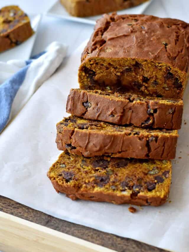 A sliced loaf of gluten free chocolate chip pumpkin bread on parchment paper with a blue and white striped kitchen towel and more slices on white plates in the background.