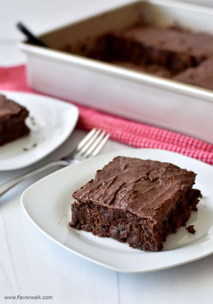 Close up view of a slice of gluten free chocolate zucchini cake on a small white plate with another serving and the rest of the cake in the background.