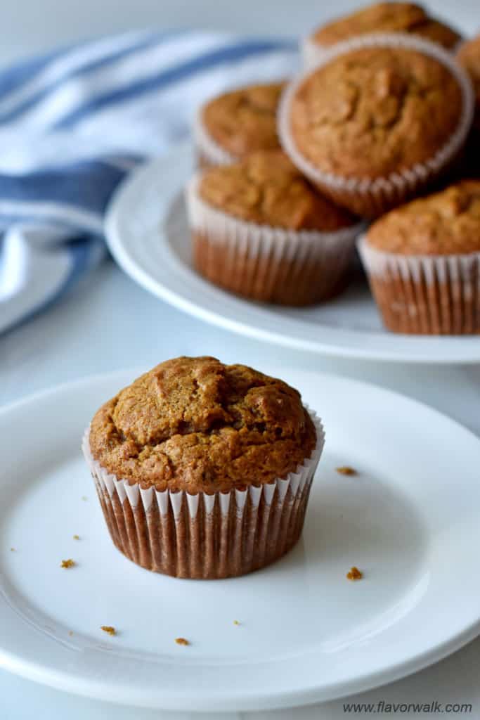 One gluten free pumpkin banana muffin on small white plate with more muffins and a blue and white striped kitchen towel in the background.