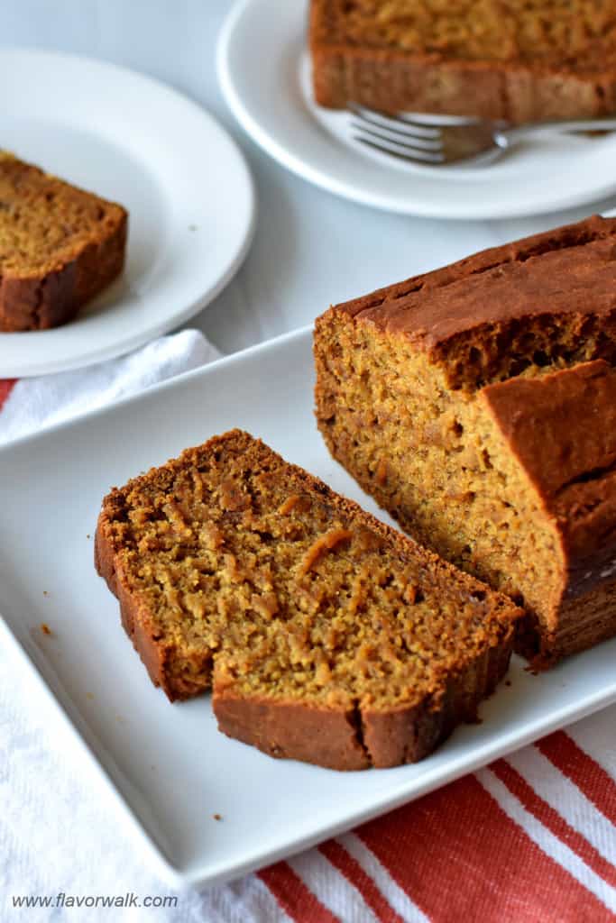 One slice and loaf of gluten free pumpkin banana bread on white serving platter with more slices of bread in the background.