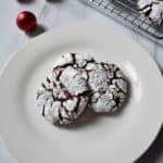 Close up of three gluten free chocolate crinkle cookies on round white plate with more cookies in the background.