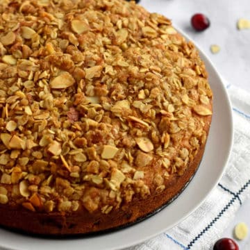 Uncut gluten free cranberry coffee cake on a round white plate with fresh cranberries and sliced almonds scattered around the plate.