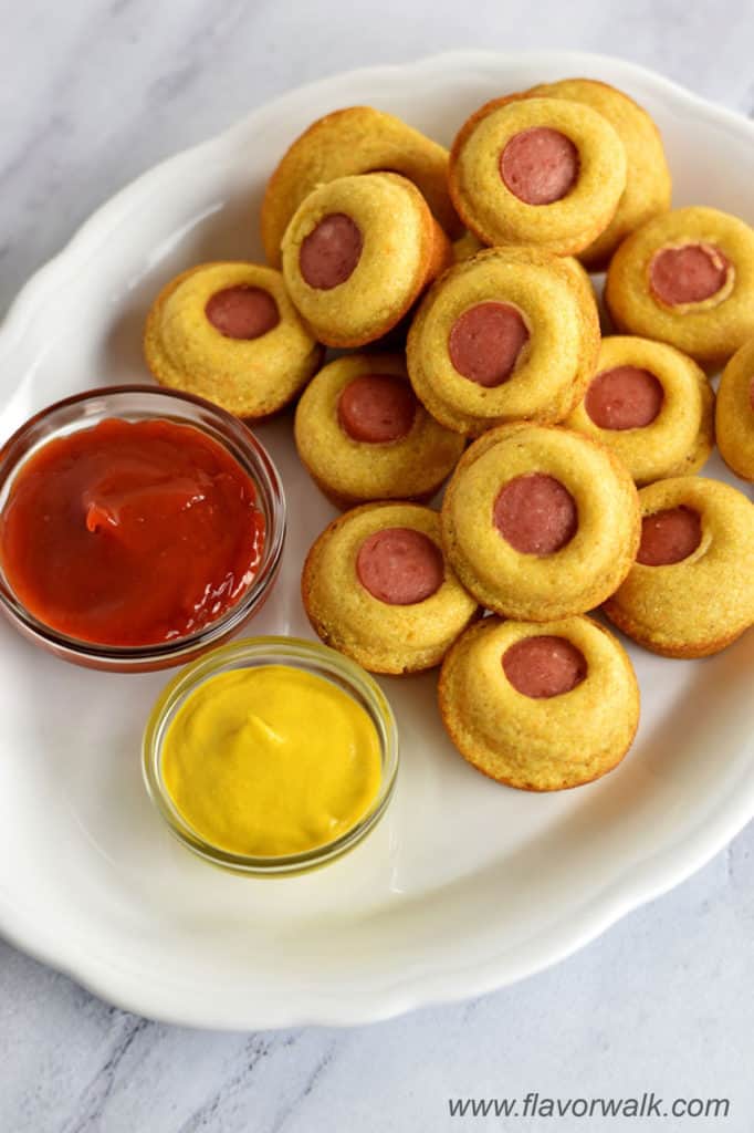 Close-up view of a white platter filled with mini corn dog muffins and small bowls containing ketchup and mustard.