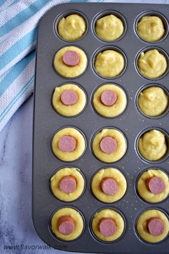 Overhead view of mini muffin pan filled with cornbread batter and a small hot dog in some of the cavities.