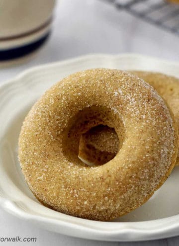Close-up of two gluten free cinnamon sugar donuts on small white plate with a coffee cup and more donuts in the background.