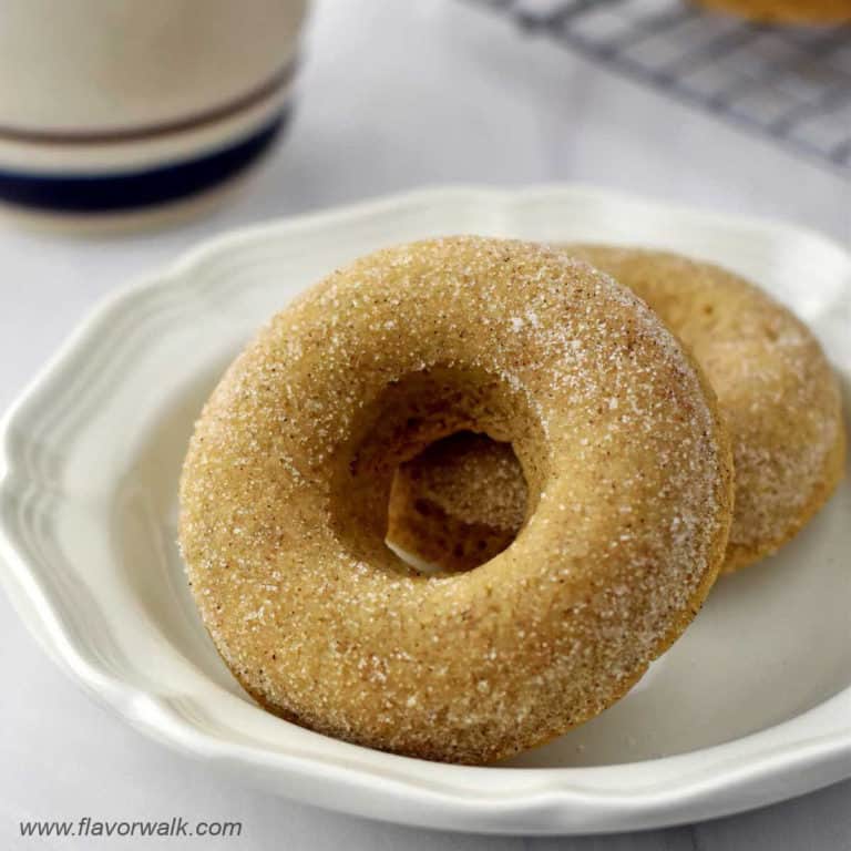 Close-up of two gluten free cinnamon sugar donuts on small white plate with a coffee cup and more donuts in the background.