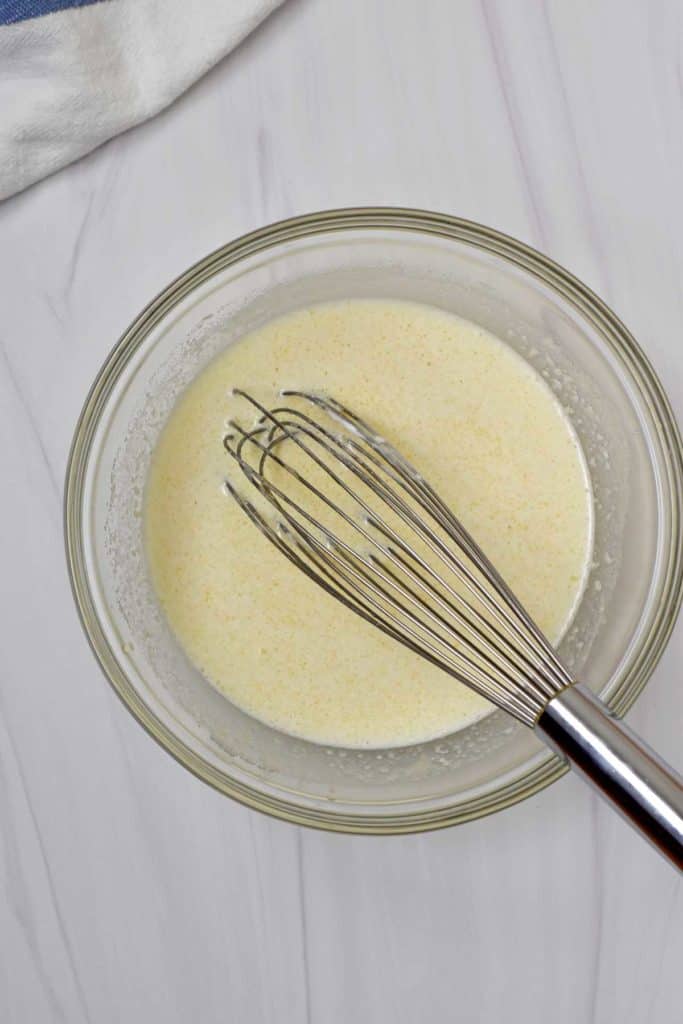 Overhead view of glass mixing bowl containing egg, sugar, vanilla, milk, sour cream, and melted butter whisked together with wire whisk.