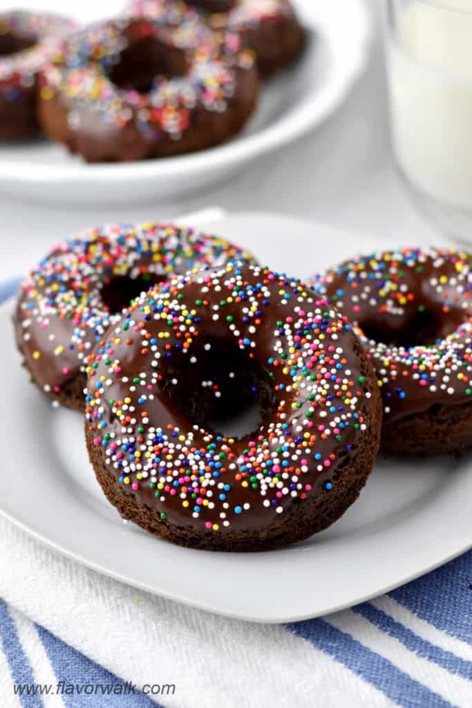 Three chocolate donuts with sprinkles on a white dessert plate with more donuts and a glass of milk in the background.