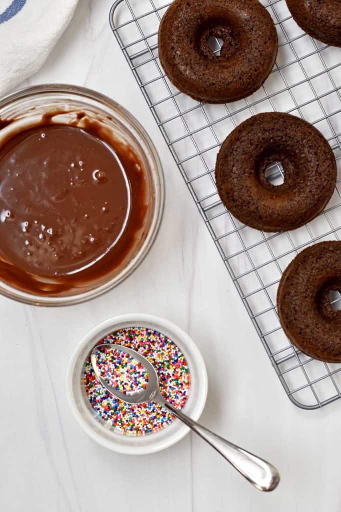Overhead view of chocolate glaze, rainbow sprinkles, and unglazed chocolate donuts on wire rack.