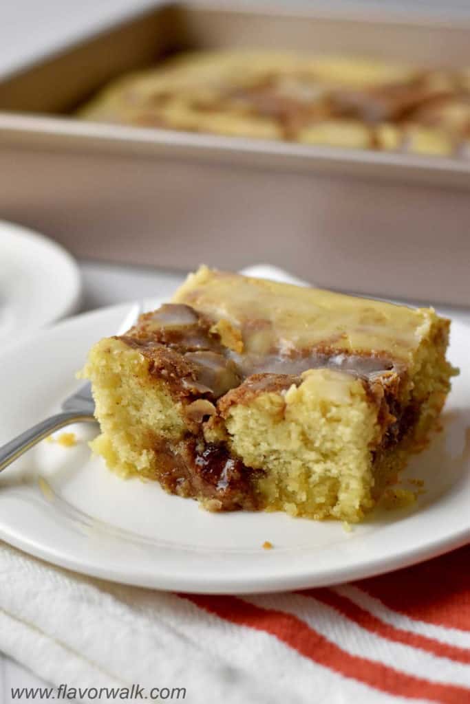 A slice of easy cinnamon roll cake and a fork on a white dessert plate with more cake in the background.