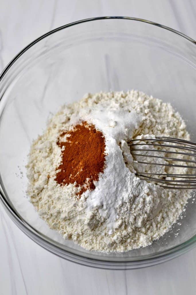 Overhead view of gluten free flour blend, salt, baking powder, baking soda, cinnamon, and a wire whisk in a glass mixing bowl.