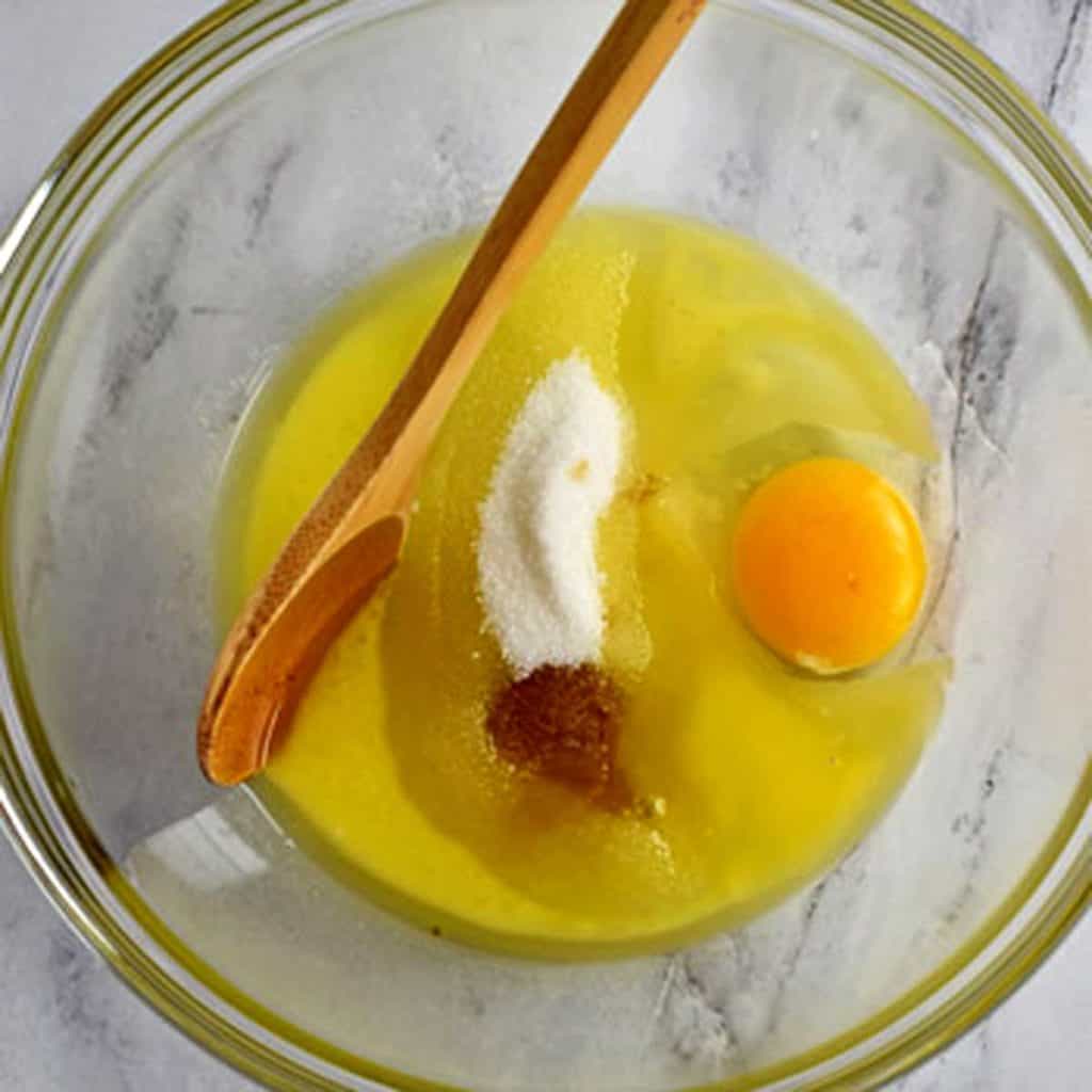 Melted butter, sugar, egg, vanilla, and wooden spoon in glass mixing bowl.