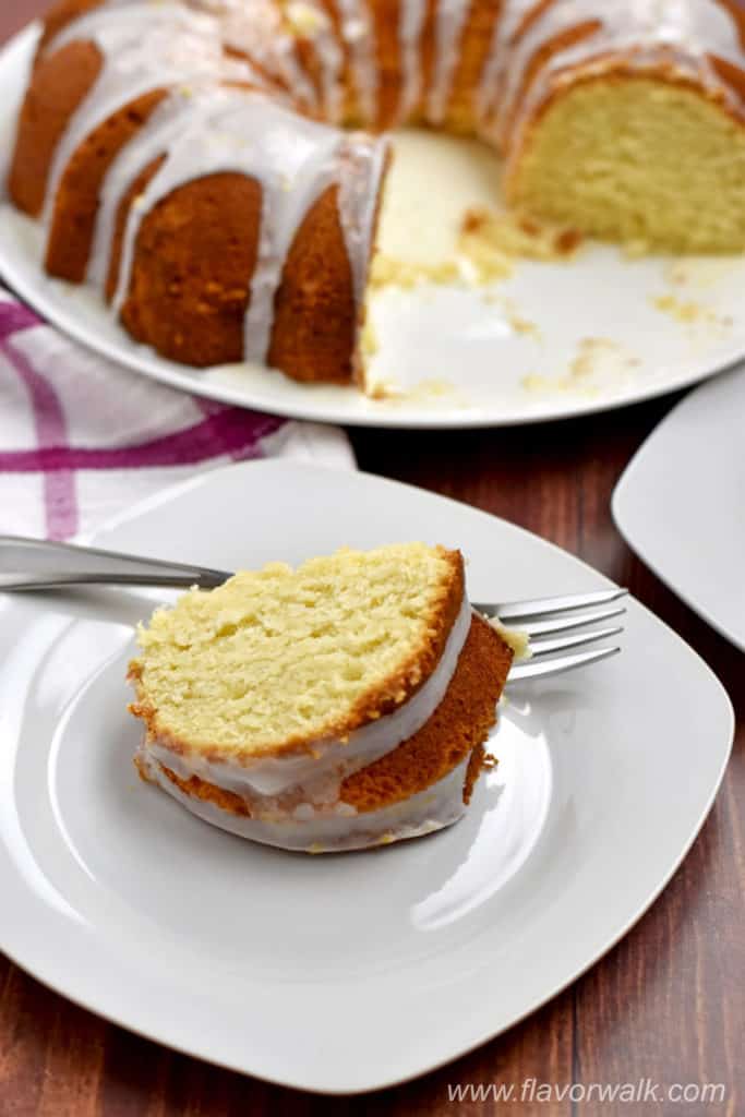 A slice of lemon cake and fork on small white plate with more cake in the background.