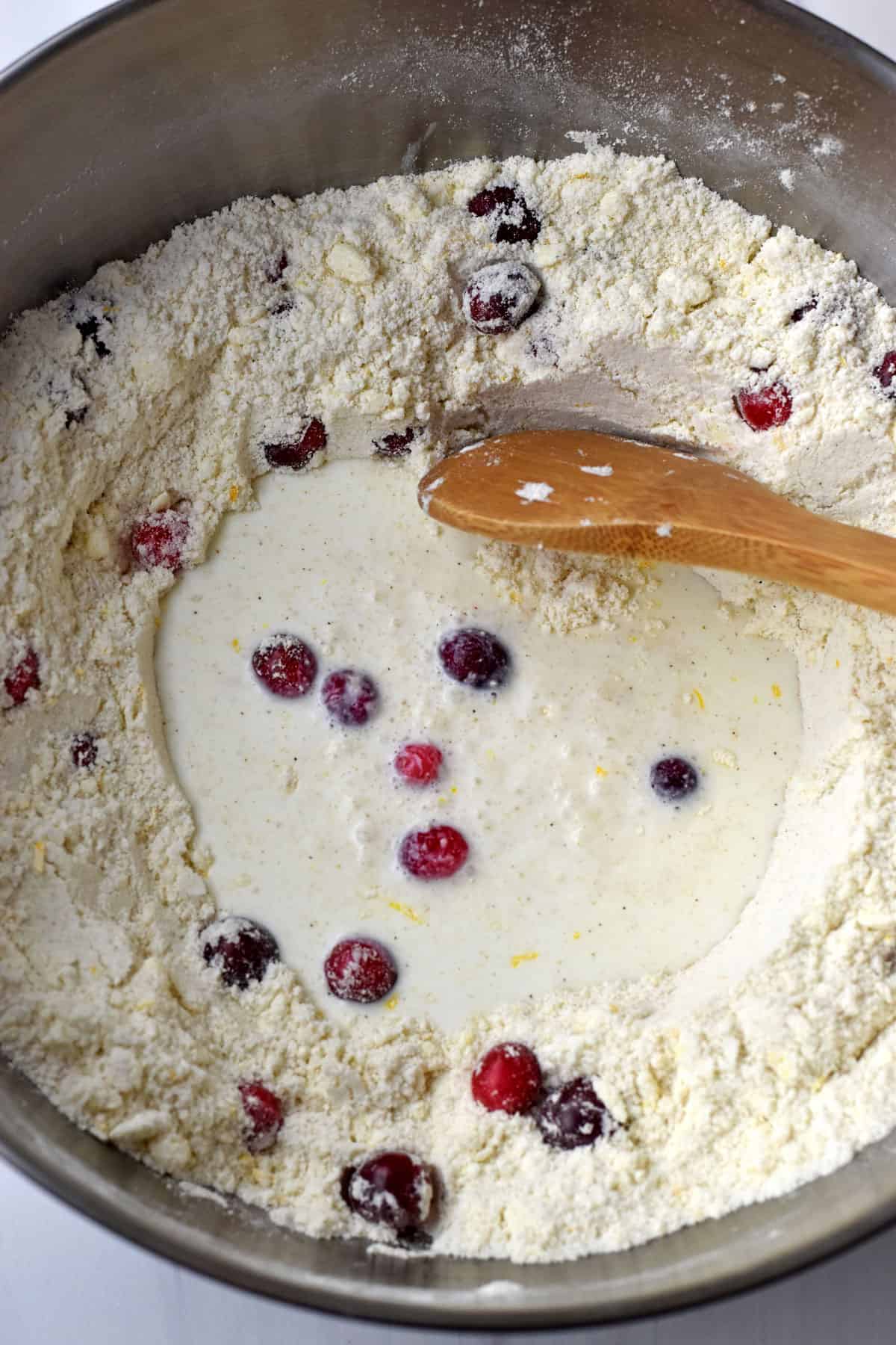 Wet ingredients and fresh cranberries being stirred into flour/butter mixture with a wooden spoon.
