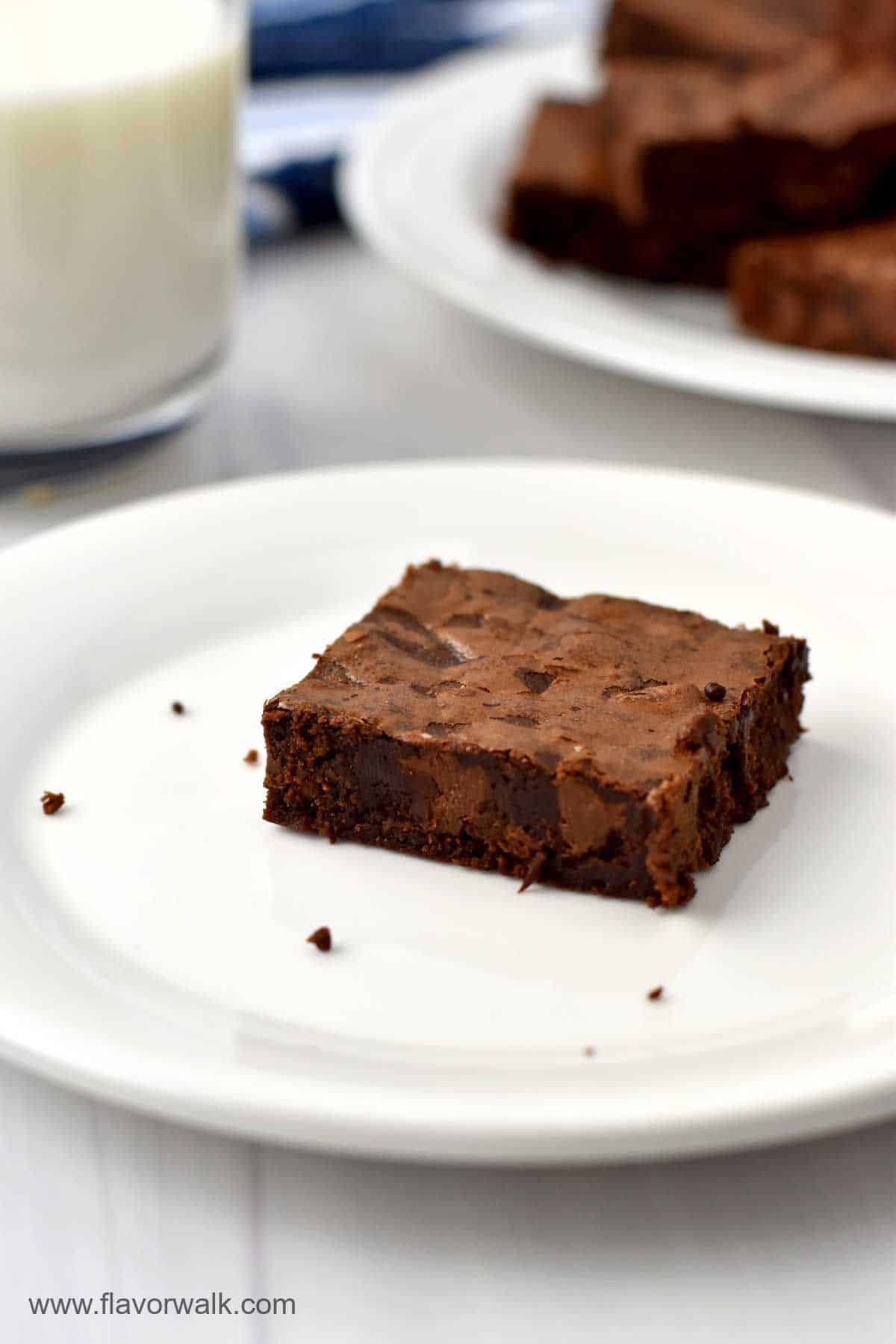 One brownie on a small white plate with a glass of milk and more brownies in the background.