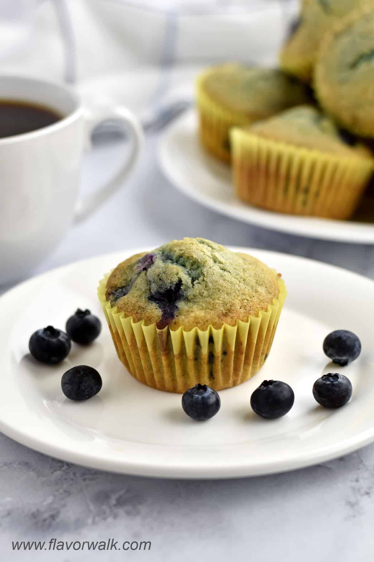 One gluten free blueberry muffin and a few blueberries on a small white plate with a cup of coffee and more muffins in the background.