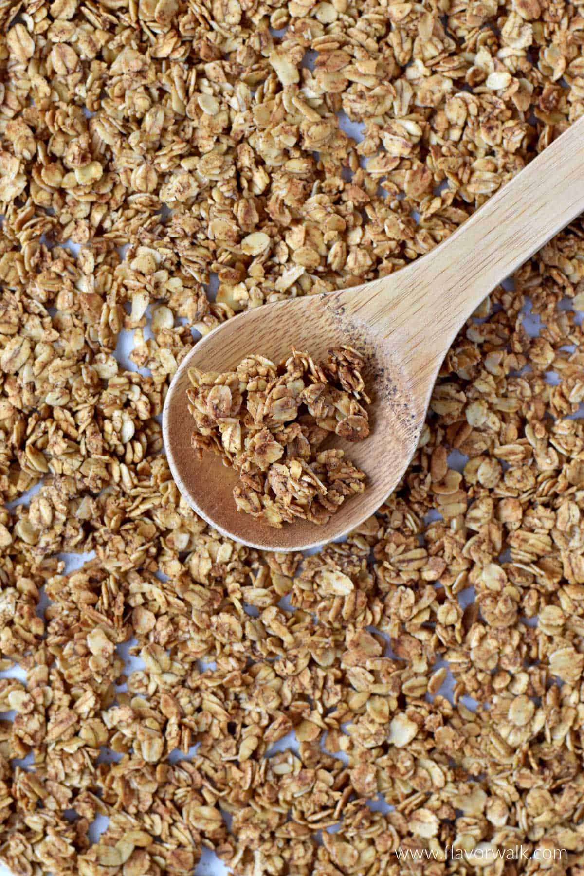 A wooden spoon with a scoop of cinnamon granola resting on a baking sheet filled with granola.