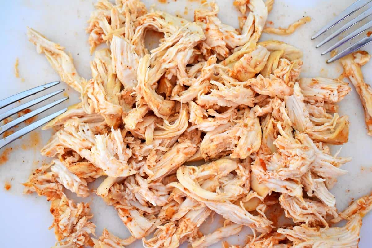 Shredded chicken and two forks on a white cutting board.