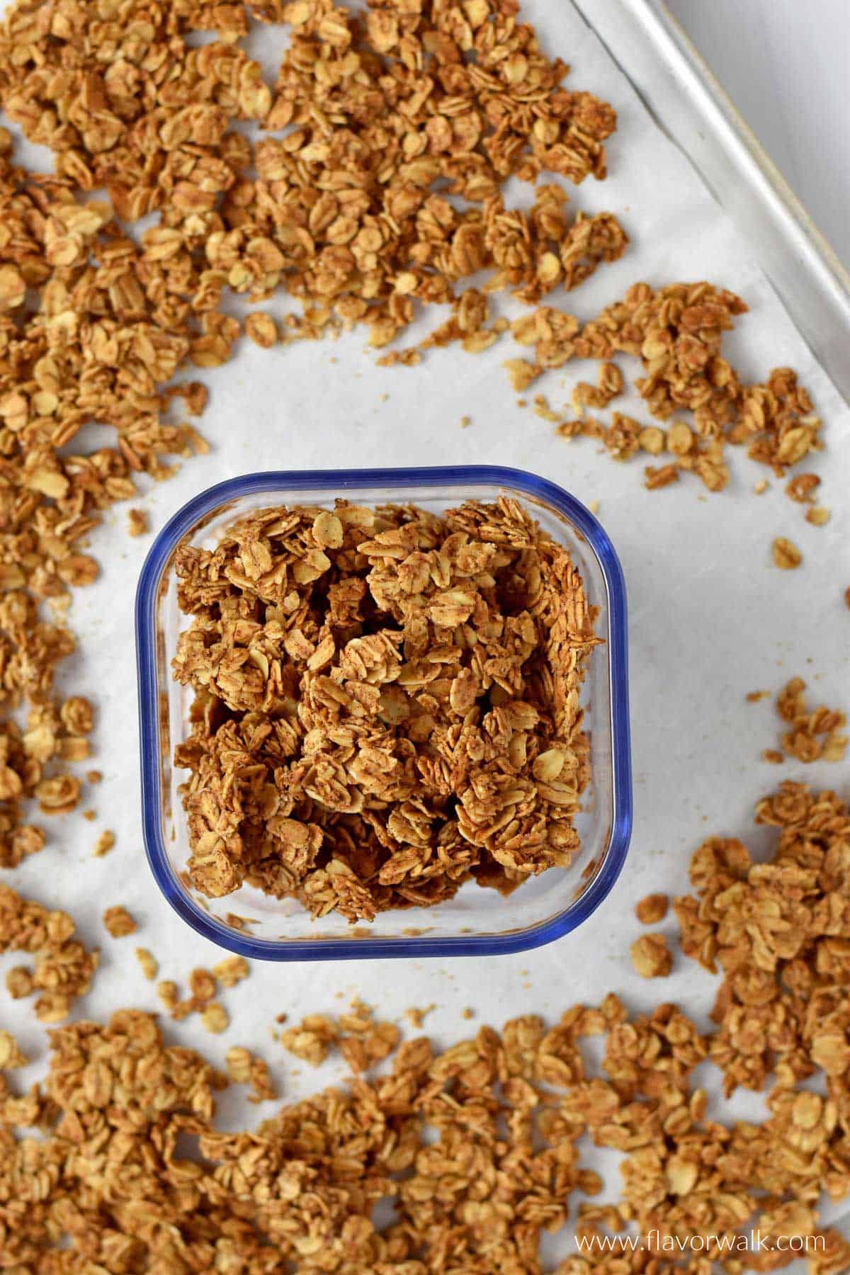 A small container with cinnamon granola on a parchment-lined baking sheet with more granola scattered around the container.