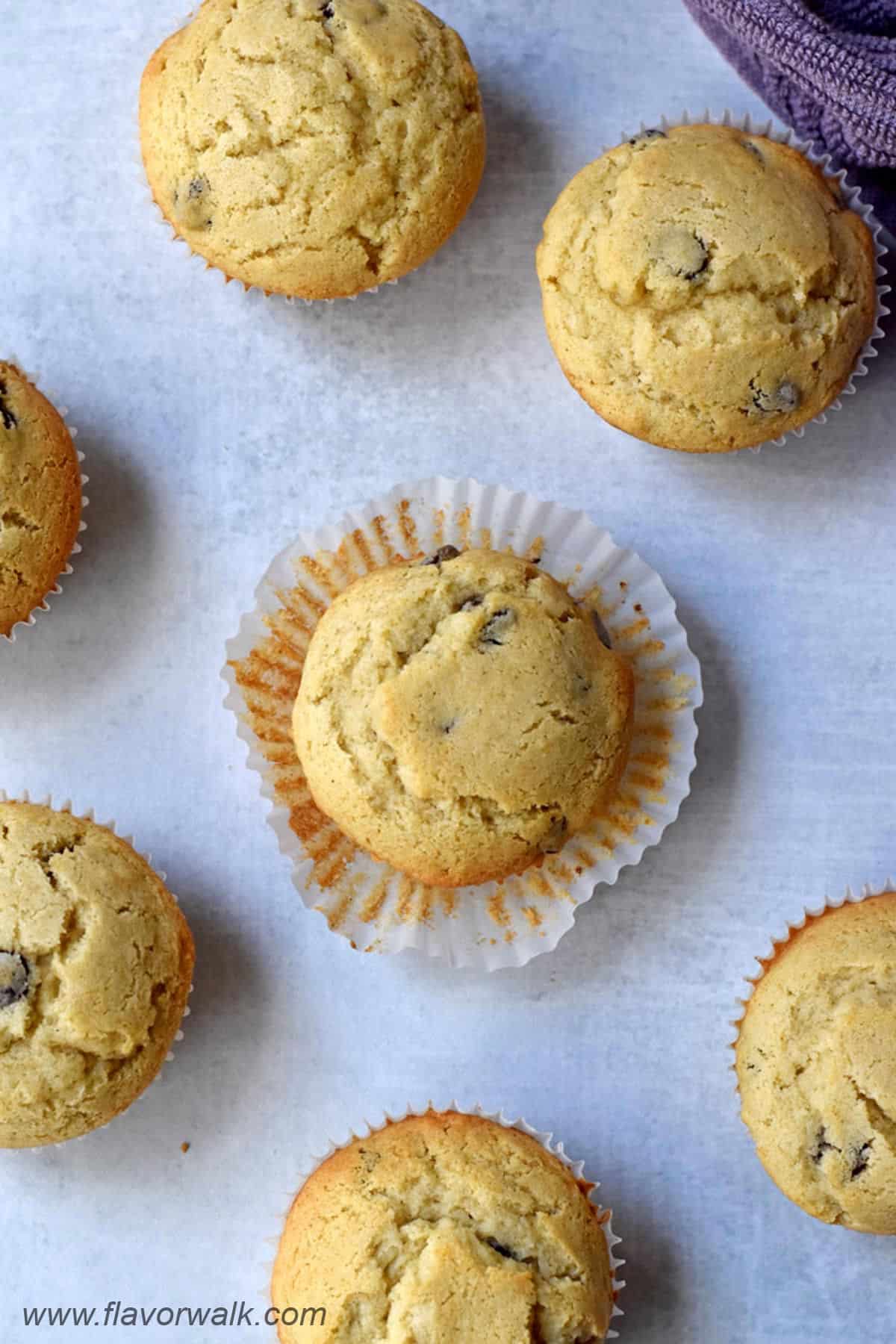 Gluten free chocolate chip muffins scattered on counter with the liner peeled from the sides of the muffin in the middle.