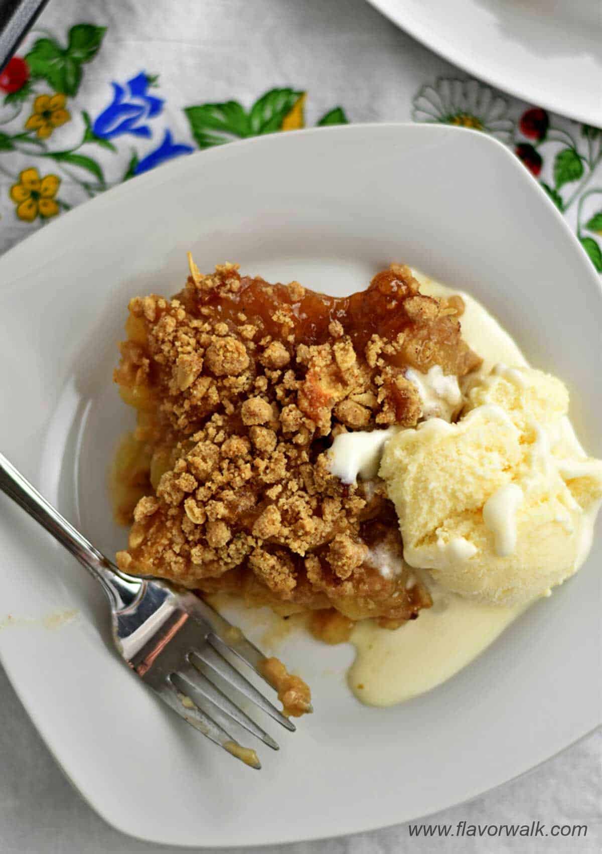 A slice of apple dessert with a scoop of vanilla ice cream and a fork on a small white plate.