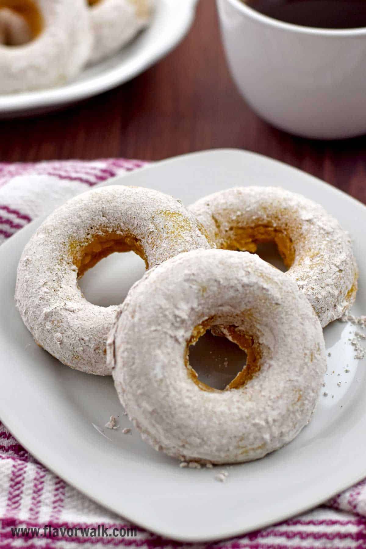 Three pumpkin donuts on a white plate with more donuts and a cup of coffee in the background.