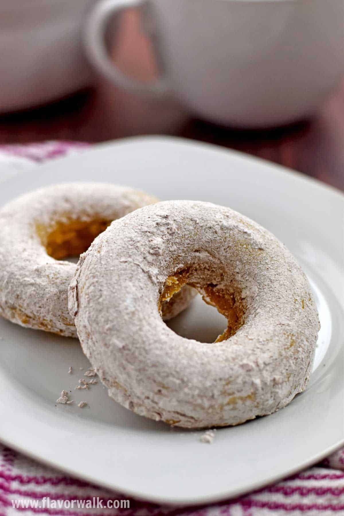 Two gluten free pumpkin donuts on a white plate with a white coffee cup in the background.