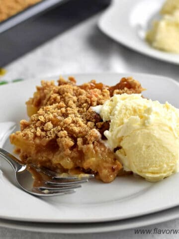 One serving of gluten free apple dessert with a scoop of ice cream and a fork on a stack of 2 white dessert plates with more apple dessert in the background.