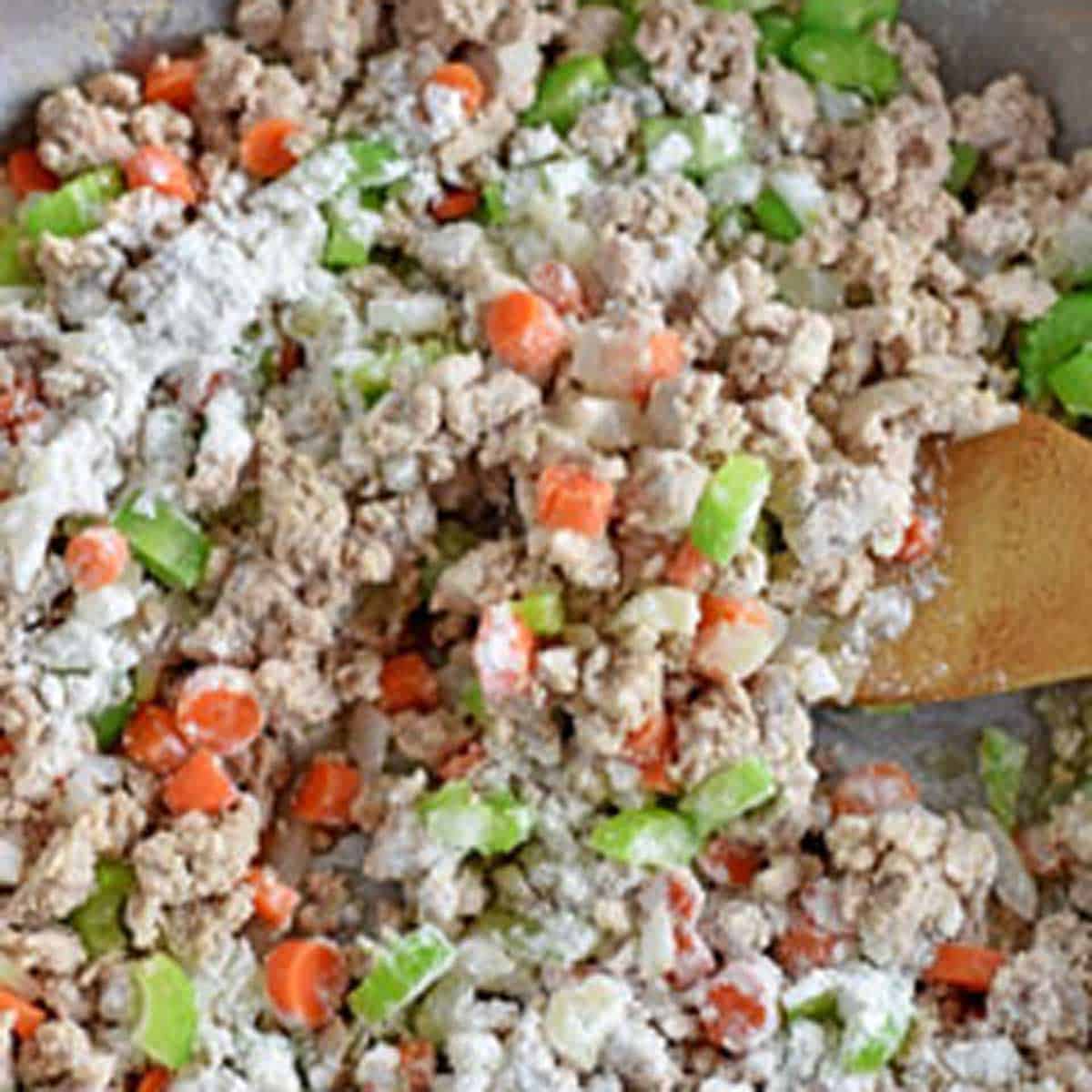 A wooden spoon stirring flour into a large skillet filled with cooked ground turkey, diced onions, celery, and carrots.