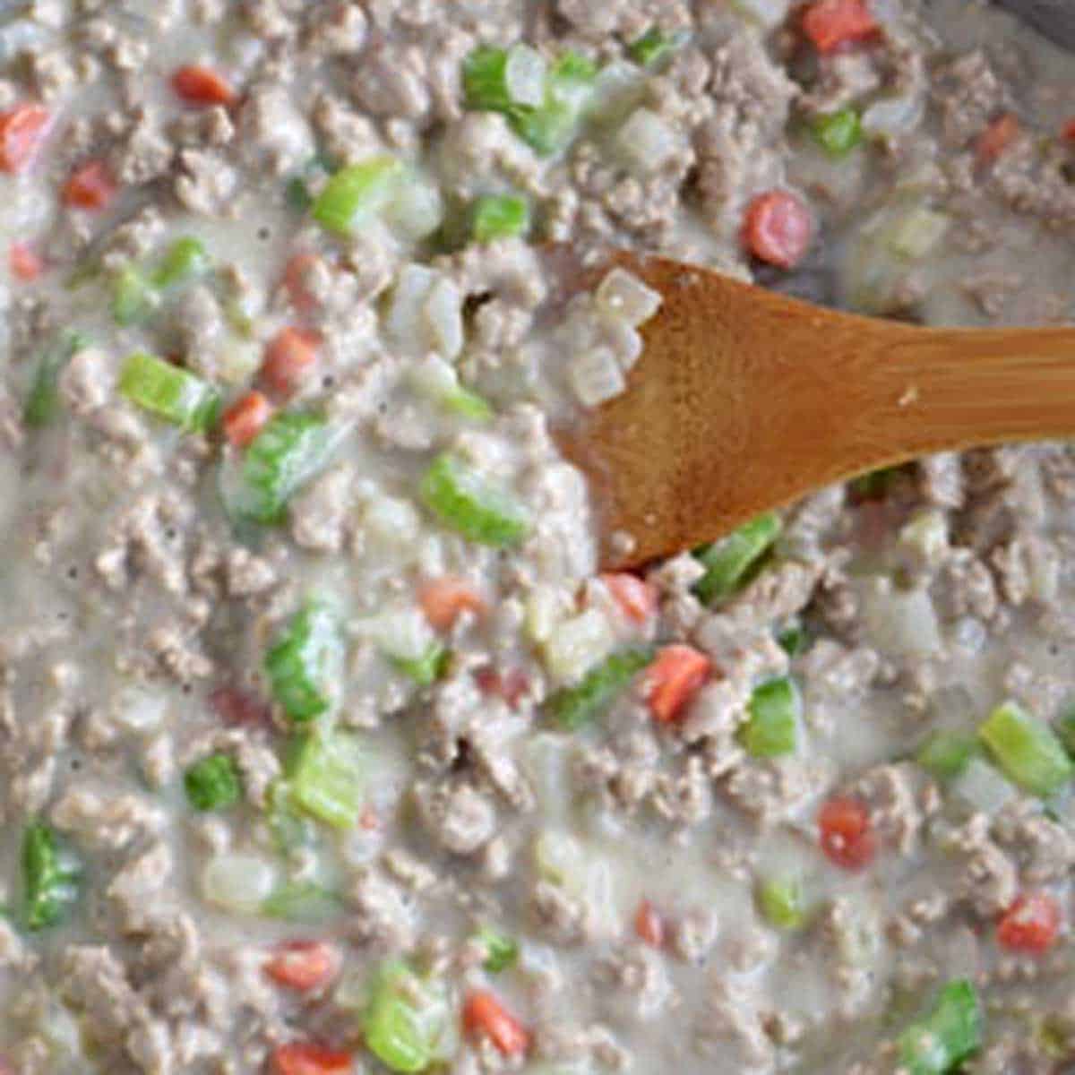A wooden spoon stirring milk into the meat and vegetables for cheeseburger soup.