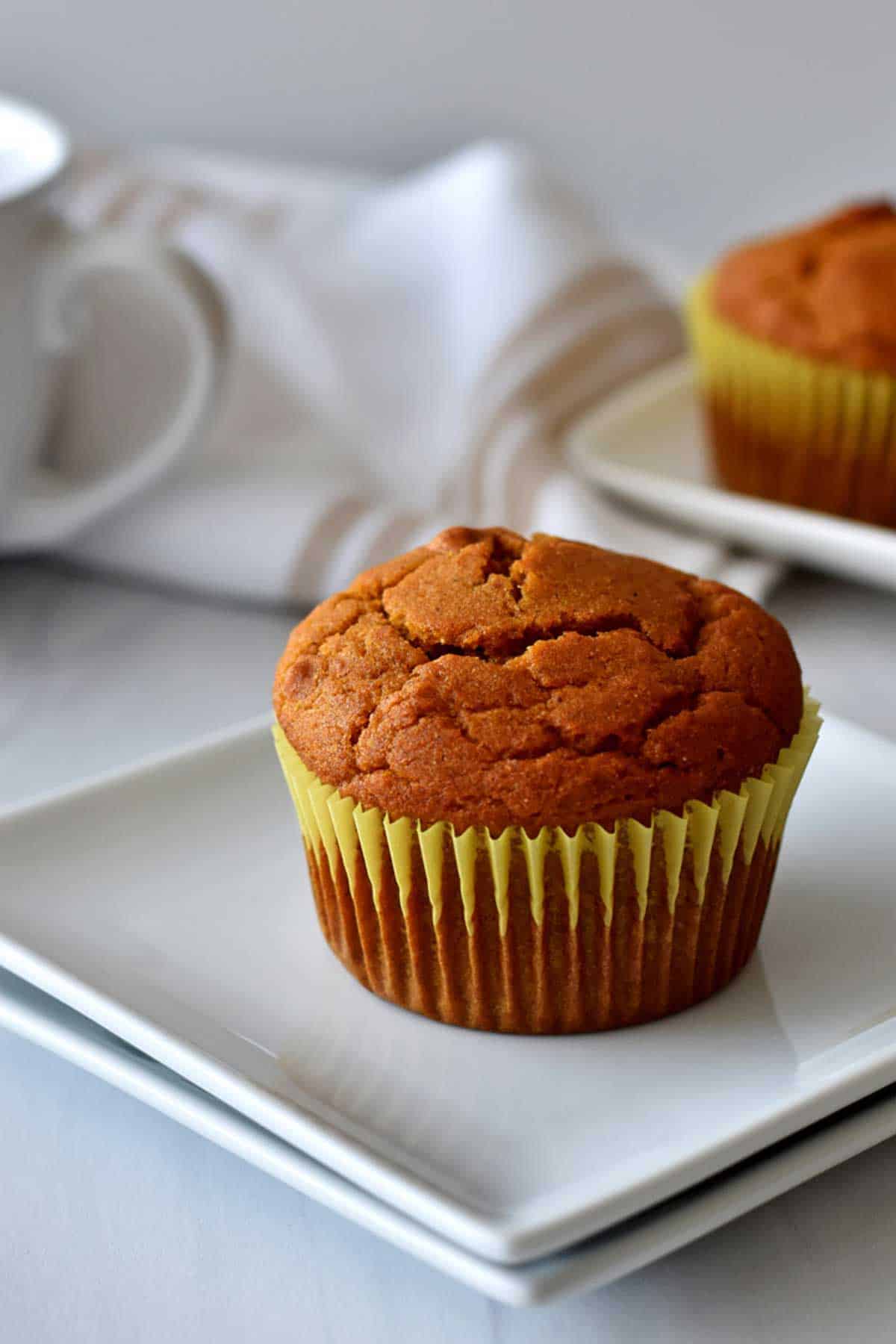 One gluten free pumpkin muffin on a stack of plates with a coffee cup, a brown and white striped towel, and another muffin in the background.