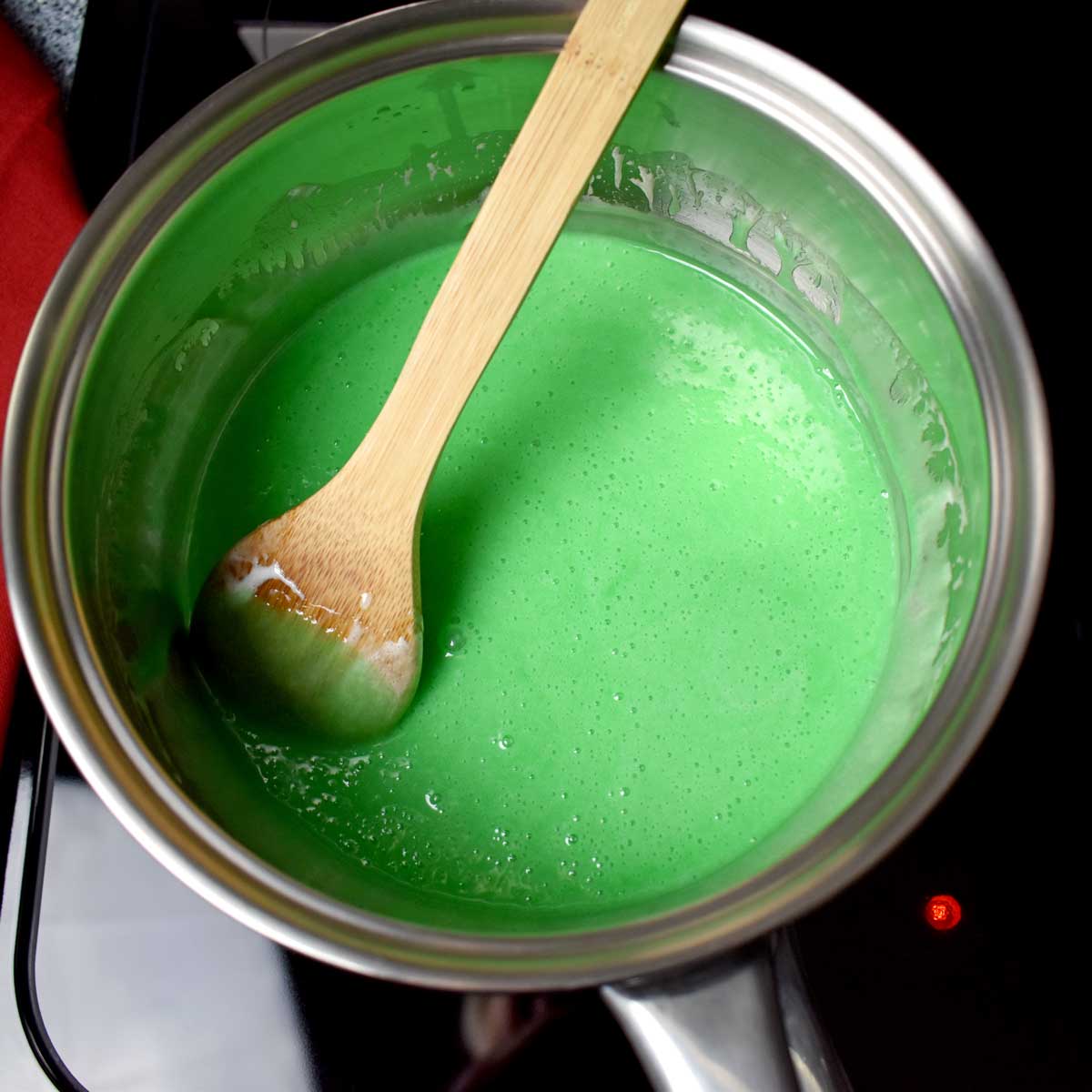 A wooden spoon and green colored marshmallow mixture for making holly cookies in large saucepan.