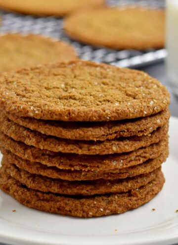 A stack of gluten free ginger snaps on a white plate with more cookies and a glass of milk in the background.