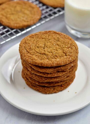 Gluten free ginger snaps stacked on a plate with more cookies and a glass of milk in the background.