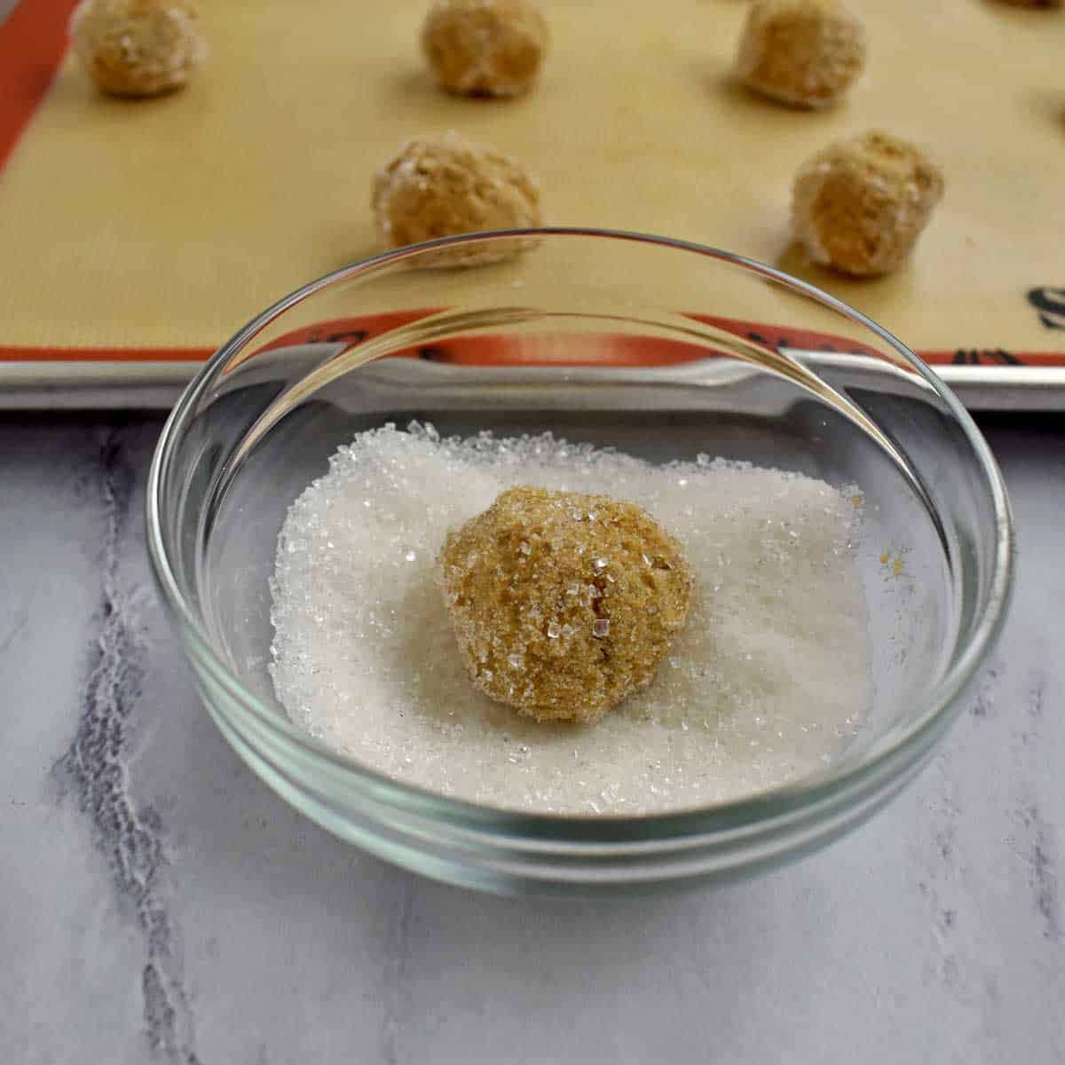 A gluten free ginger snaps dough ball in a small bowl of sugar with more dough balls on a baking pan in the background.