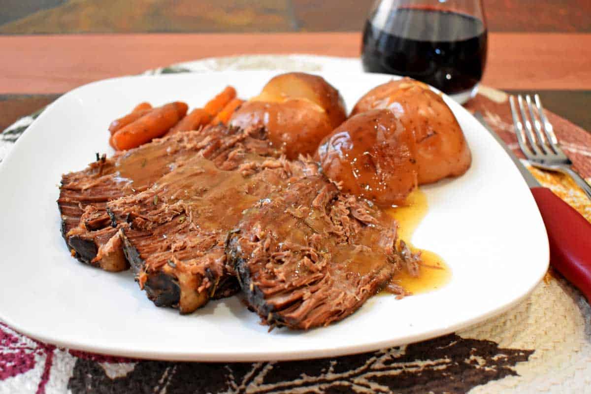 A serving of slow cooker beef pot roast and vegetables on a white dinner plate with a glass of red wine.