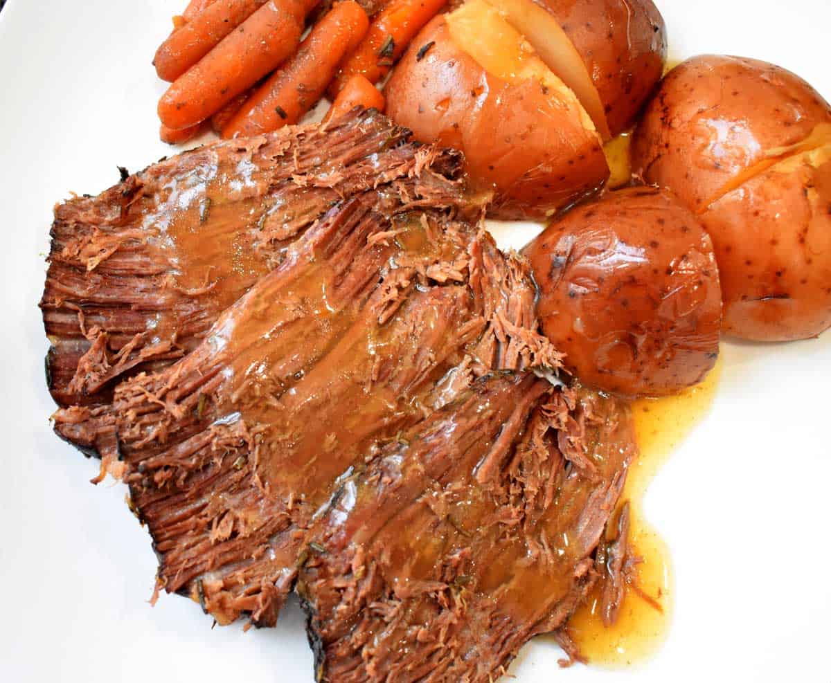 Sliced pot roast with gravy, red potatoes, and carrots on a white plate.
