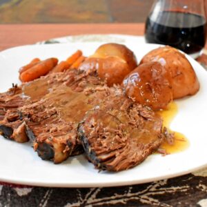 A serving of beef pot roast and gravy, red potatoes, and baby carrots on a white dinner plate with a glass of red wine in the background.