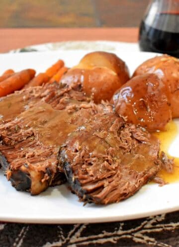 A serving of beef pot roast and gravy, red potatoes, and baby carrots on a white dinner plate with a glass of red wine in the background.