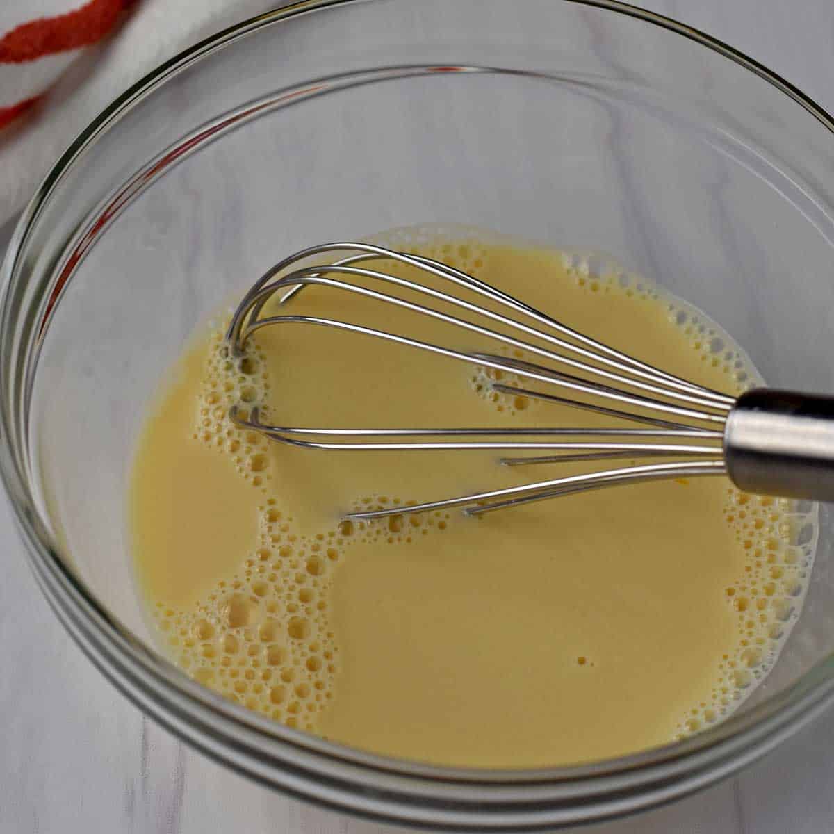 Wet ingredients for apple fritters whisked together with a wire whisk in a glass mixing bowl.