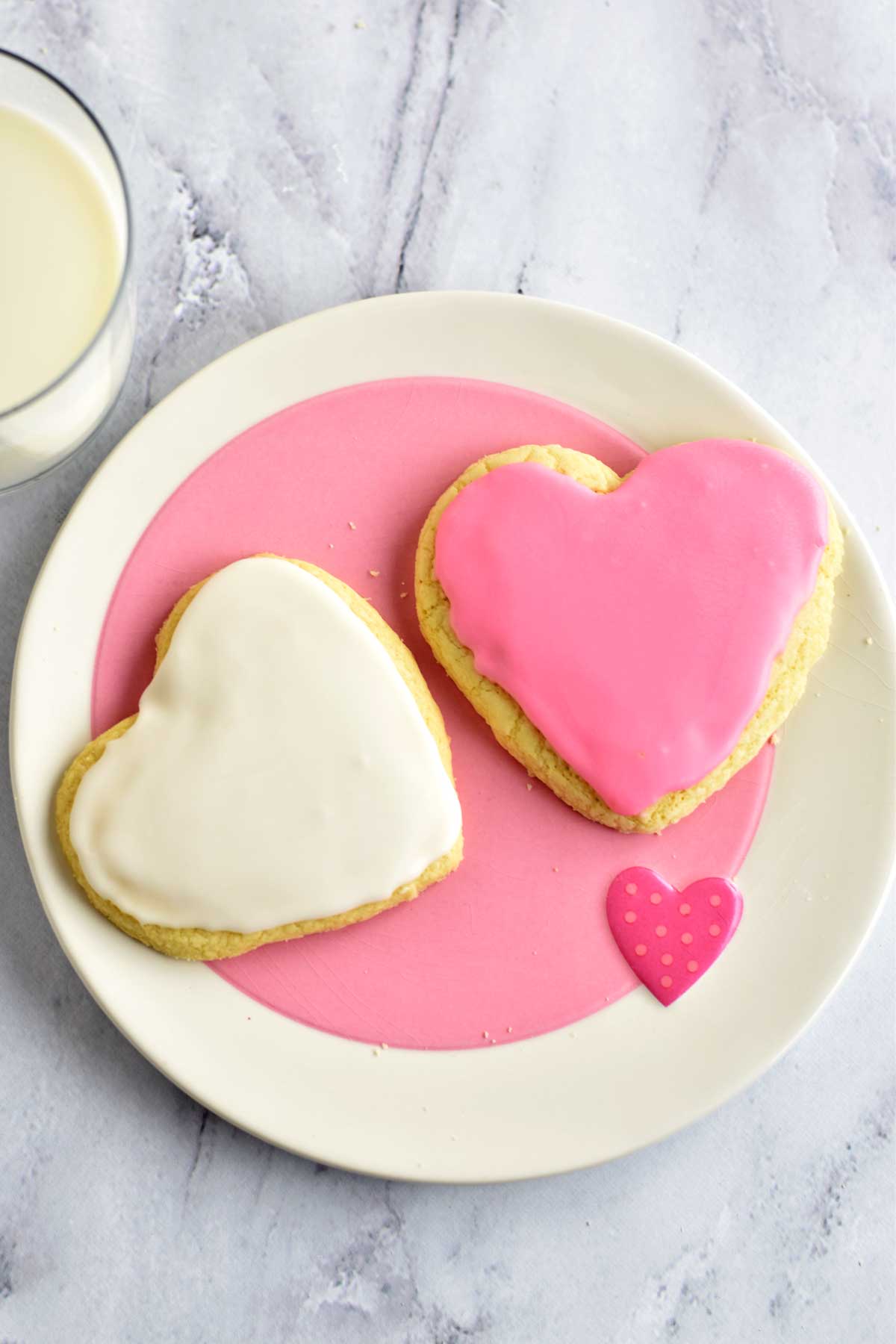 Two heart-shaped gluten free frosted sugar cookies on a pink and white plate.