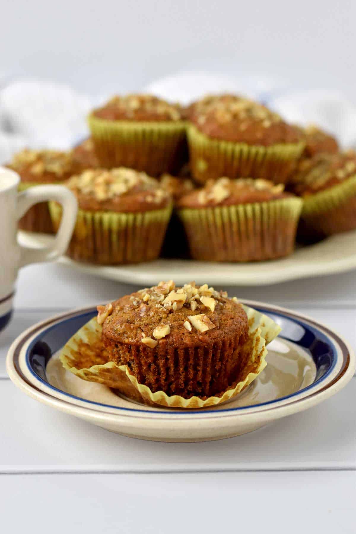 One gluten free banana nut muffin on a small plate with a coffee cup and more muffins in the background.